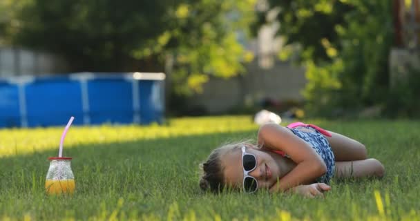 Nastoletnia dziewczyna w stroju kąpielowym i okularach przeciwsłonecznych kłamie i bawi się na zielonym trawniku podczas wakacji na tle basenu. sok ze słomką w słoiku. Aktywność wakacyjna — Wideo stockowe