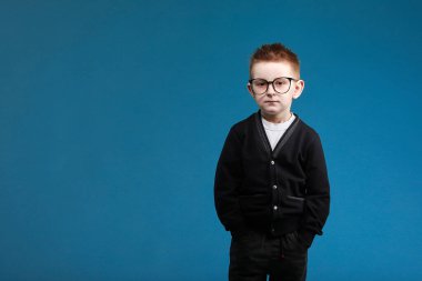 Okula dönelim. İnek gözlüklü ve okul üniformalı sevimli çocuk mavi arka planda dururken kameraya bakıyor..