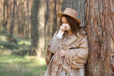 Güneşli bir günde parkta öksüren koruyucu yüzlü, şapkalı ve tıbbi maskeli genç bir kadının portresi. Virüs, gribi önlemek için karantinaya alınıyor.