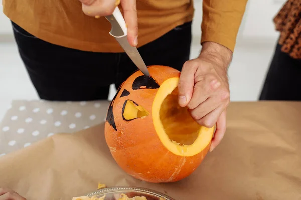 Close up of man hands carving Jack O Lattern from ripe orange pumpkin with knife on her wooden kitchen table. male preparing all hallows eve Halloween party decorations. Background, copy space