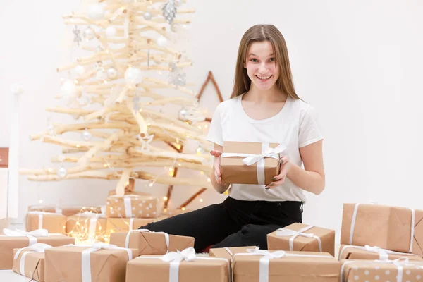 Nochebuena. Linda chica se sienta cerca del árbol de Navidad con un montón de cajas de regalo. La casa está decorada con vacaciones. Bajo el árbol de Navidad hay muchos regalos. Chica es feliz y sonriente —  Fotos de Stock