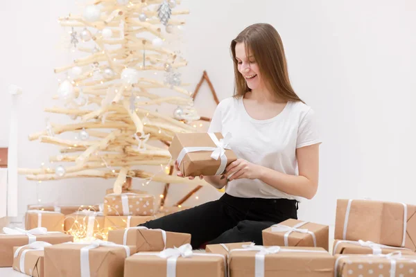 Nochebuena. Linda chica se sienta cerca del árbol de Navidad con un montón de cajas de regalo. La casa está decorada con vacaciones. Bajo el árbol de Navidad hay muchos regalos. Chica es feliz y sonriente —  Fotos de Stock