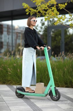 Elektrikli scooter kullanan genç mutlu bir kadın yaz günü iş alanında şehir sokağında karton kutular taşıyor. Sürdürülebilirlik, dağıtım ve çevre dostu modern yaşam tarzı kavramı
