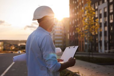 Erkek mimarın yan görünüşü, inşaat mühendisi güvenlik incelemesi modern konut inşaatının yeni projesini inceliyor. İnşaat mühendisi notlar ve günbatımı ışınları için planlar ve pano tutuyor.