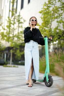 Elektrikli scooter 'ın yanında güneş gözlüklü neşeli bir kadın ya da yazları modern yerleşim bölgesinde akıllı telefondan e-scooter konuşmaları. Kentsel çevre dostu yaşam tarzı ve sürdürülebilirlik kavramı