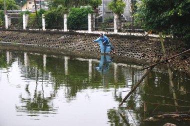 Hanoi, Vietnam: 30 Aralık 2017 - Mavi yağmurluk giyen kimliği belirsiz bir balıkçı yağmurlu bir günde balıkçı oltasını şehir gölüne attı. Göl demir bir çitle çevrili ve beton takviyeli bir kıyıyla çevrili..