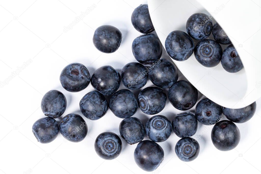Berries of ripe juicy blueberries are scattered on from a white ceramic cup on white, top view, place for text