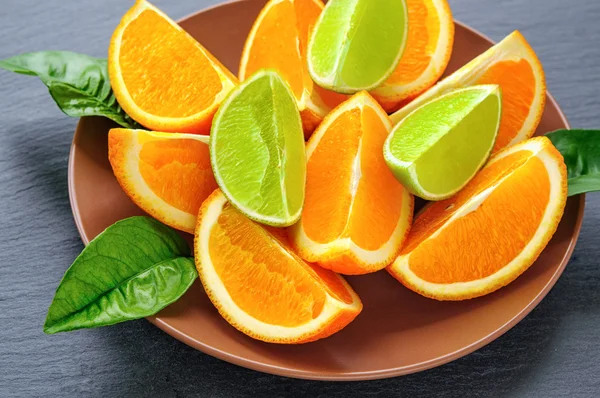 Orange and lime sliced segments  with green leaves on brown plate, black slate stone. Vitamin concept — Stock Photo, Image