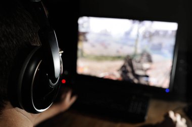 Karanlık odada kulaklıklarla video oyunu oynayan genç bir adamın yan görüntüsü. Ön planda siyah kulaklıklar, arka planda bulanık bir bilgisayar ekranı. Sanal spor oyuncusu.