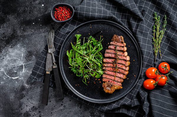 Sliced sirloin steak, marbled beef meat with arugula. Black background. Top view.