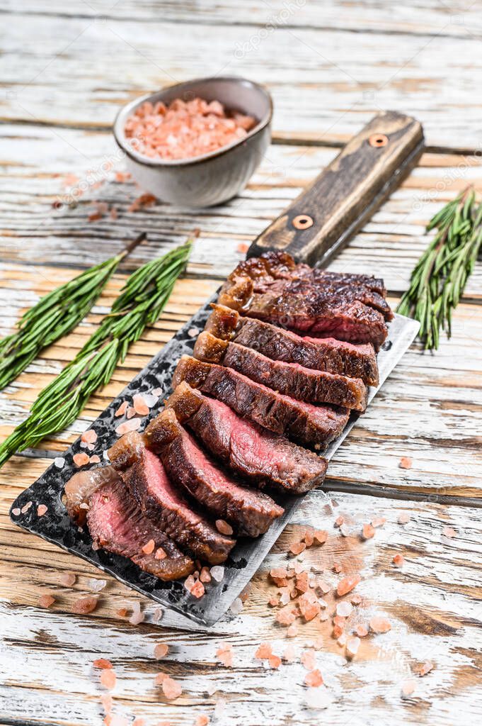 Grilled top sirloin cap or picanha steak on a meat cleaver with herbs. White wooden background. Top view.