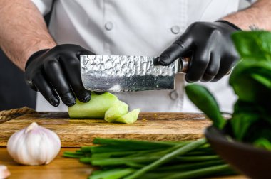 Siyah eldivenli bir şef, ahşap bir tahtanın üzerinde taze yeşil bir salatalık kesiyor. Sağlıklı organik gıda pişirme kavramı.