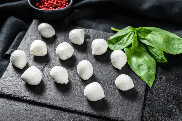 Üzerinde fesleğen yaprağı ve pembe biber olan taş bir tahtada mini mozzarella peyniri topları. Siyah arka plan. Üst görünüm.
