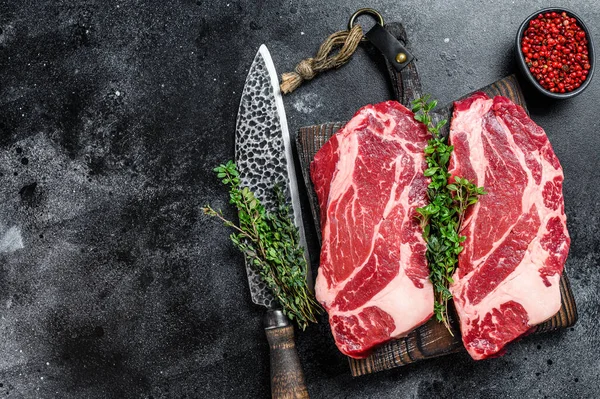 Raw chuck eye roll steaks premium beef meat on a cutting board. Black background. Top view. Copy space.