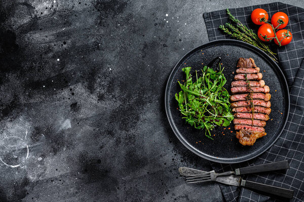 Cooked grilled strip loin steak, marbled beef meat with arugula. Black background. Top view. Copy space