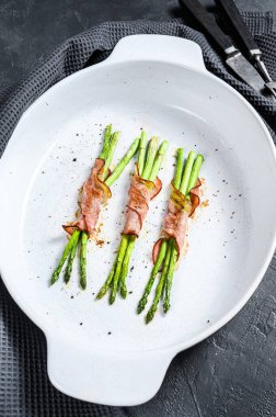 Pişirme kabında kuşkonmaz etrafında lezzetli domuz pastırması. Siyah arka plan. Üst görünüm.