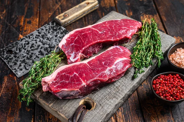 Raw sirloin beef meat steak on a wooden cutting board with herbs. Dark wooden background. Top view.