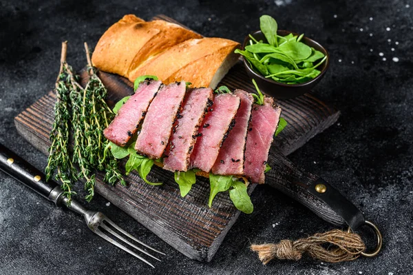 Baguette tonhal steak szendvics arugulával a vágódeszkán. Fekete háttér. Felülnézet — Stock Fotó