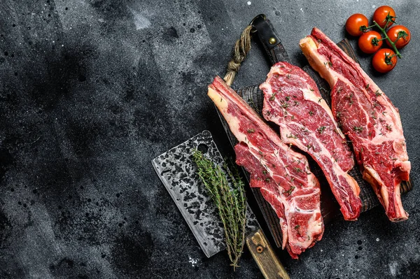 Assortment of raw cuts beef meat steaks on the bone. Black background. Top view. Copy space.