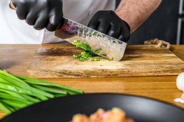 Siyah eldivenli bir şef, ahşap kesim tahtasında taze yeşil soğan dilimliyor..