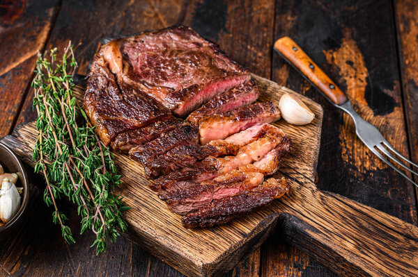 Roasted and sliced rib eye beef meat steak on a wooden cutting board with thyme. Dark wooden background. Top view.