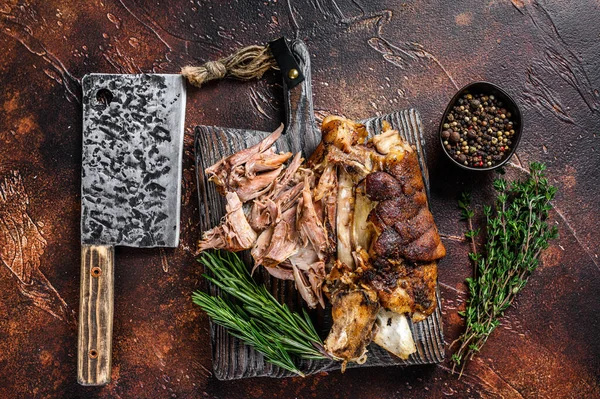 Roasted and cut German pork knuckle eisbein meat on a wooden board with meat cleaver. Dark background. Top view