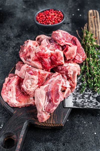 Raw lamb meat stew cuts with bone on wooden butcher board and cleaver. Black background. Top view