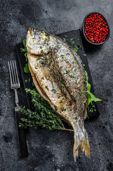 Roasted sea bream fish with herbs on a cutting board. Black background. Top view