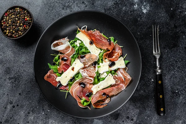 Salada de presunto de crudo de prosciutto com queijo de camembert de brie e arugula em uma chapa. Fundo preto. Vista superior — Fotografia de Stock