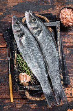 Tahta tepside otlu deniz levreği ya da çiğ balık. Koyu ahşap arka plan. Üst görünüm