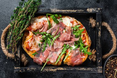 İtalyan pizzası jambonlu jambon, roka salatası ve rustik ahşap tepside peynir. Siyah arka plan. Üst görünüm