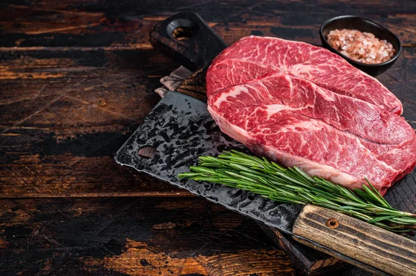 Uncooked Raw Shoulder Top Blade or flat iron beef meat steaks on a wooden butcher board with meat cleaver. Dark wooden background. Top View. Copy space