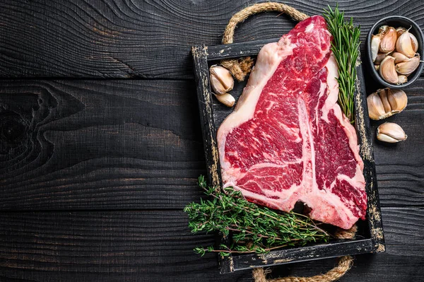 Porterhouse nebo T-bone syrové hovězí maso Steak s bylinkami a solí v dřevěném podnosu. Černé dřevěné pozadí. Horní pohled. Kopírovat prostor — Stock fotografie