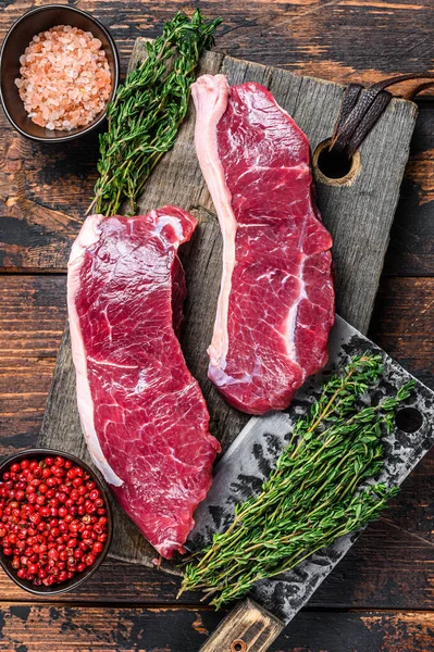 Raw sirloin beef meat steak on a wooden cutting board with herbs. Dark wooden background. Top view