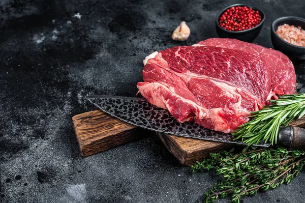 Raw Chuck eye roll Black Angus prime beef steak on butcher board with knife. Black background. Top view. Copy space