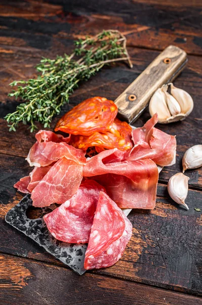 Spanish cured meat salami, jamon, choriso cured sausages on a meat cleaver. Dark wooden background. Top view