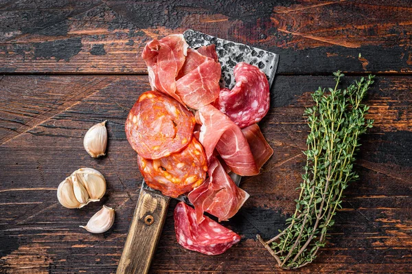 Spanish cured meat salami, jamon, choriso cured sausages on a meat cleaver. Dark wooden background. Top view