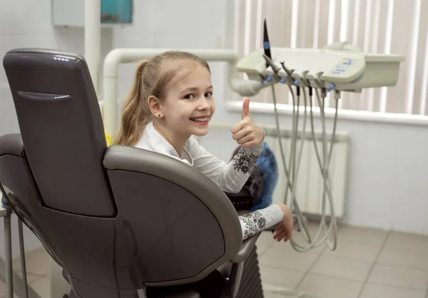 A smiling girl sits in a dental chair and gives a thumbs-up.