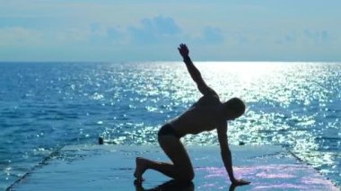 Kaslı vücutlu yakışıklı bir adam denizin arka planında esneme egzersizleri yapıyor. Sporcu yoga pozisyonunda duruyor. Yavaş çekim.