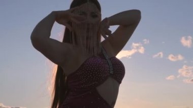 Gün batımında, başında mücevherlerle göbek dansı yapan orta boy bir kadın. Güzel bir kadın kameraya bakar..