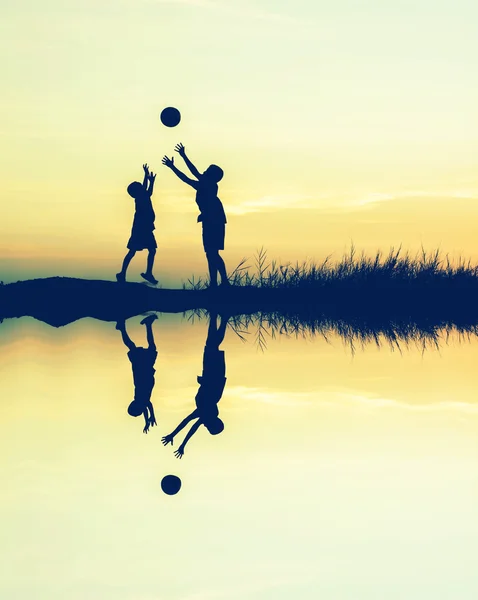Хлопчики грають у футбол на заході сонця з відображенням води. силует — стокове фото