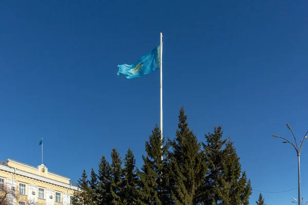 Kazakistan bayrağı ağaçların arkasındaki binanın yanındaki bayrak direğinde asılı.