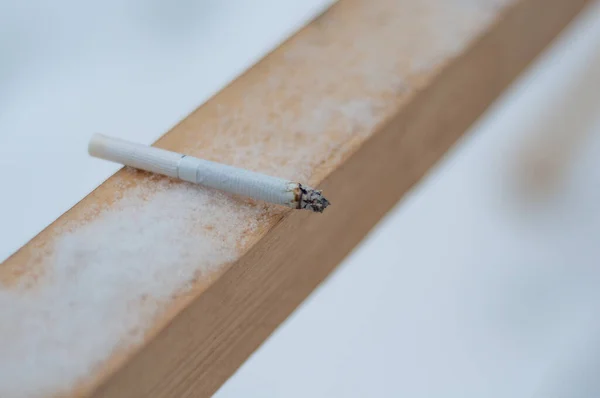 a lit cigarette with a white filter lies on a wooden railing against a background of snow