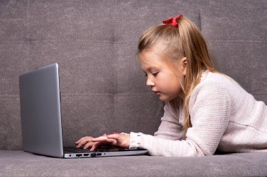 Küçük beyaz kız gri bir kanepede yatarken dizüstü bilgisayarla meşgul.