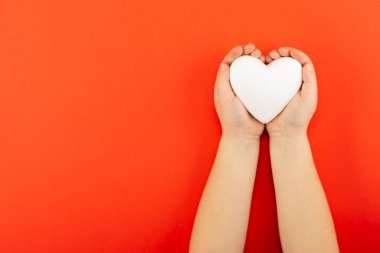 Kırmızı arka planda avuçlarında beyaz kalp taşıyan çocuk, aşk ve sağlık kavramı.