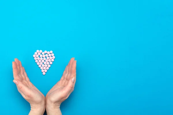 Pink pills in the shape of a heart on a blue background and female hands surround a heart made of pills.