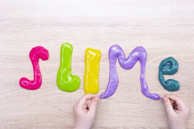 Tahta bir masanın üzerindeki çok renkli balçıktan yapılmış balçık kelimesi. Ahşap bir yüzeydeki renkli balçıktan yapılmış sümüksü yazı. Childs elleri ve balçık