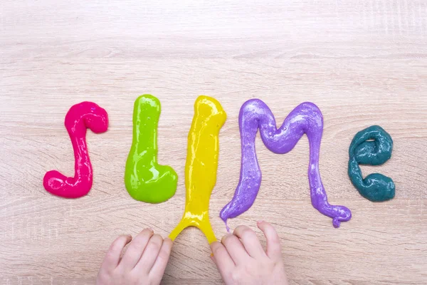 Tahta bir masanın üzerindeki çok renkli balçıktan yapılmış balçık kelimesi. Ahşap bir yüzeydeki renkli balçıktan yapılmış sümüksü yazı. Çocuk elleri balçıktan bir yazı yapar..