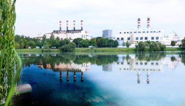Metal bir boru hattından dökülen kanalizasyon borusu, kıyıda yeşil çimenler bulunan doğal bir gölete, endüstriyel bir işletmenin egzoz borularının arka planına doğru boşalıyor..