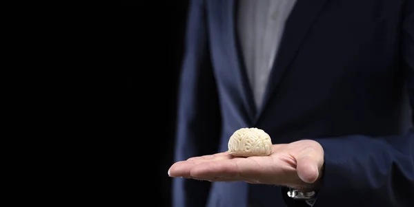 A businessman holds in his hand a model of a brain made of polymer clay on a dark background.